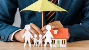 Male hands with paper cut family, house model and umbrella on wooden table, life insurance